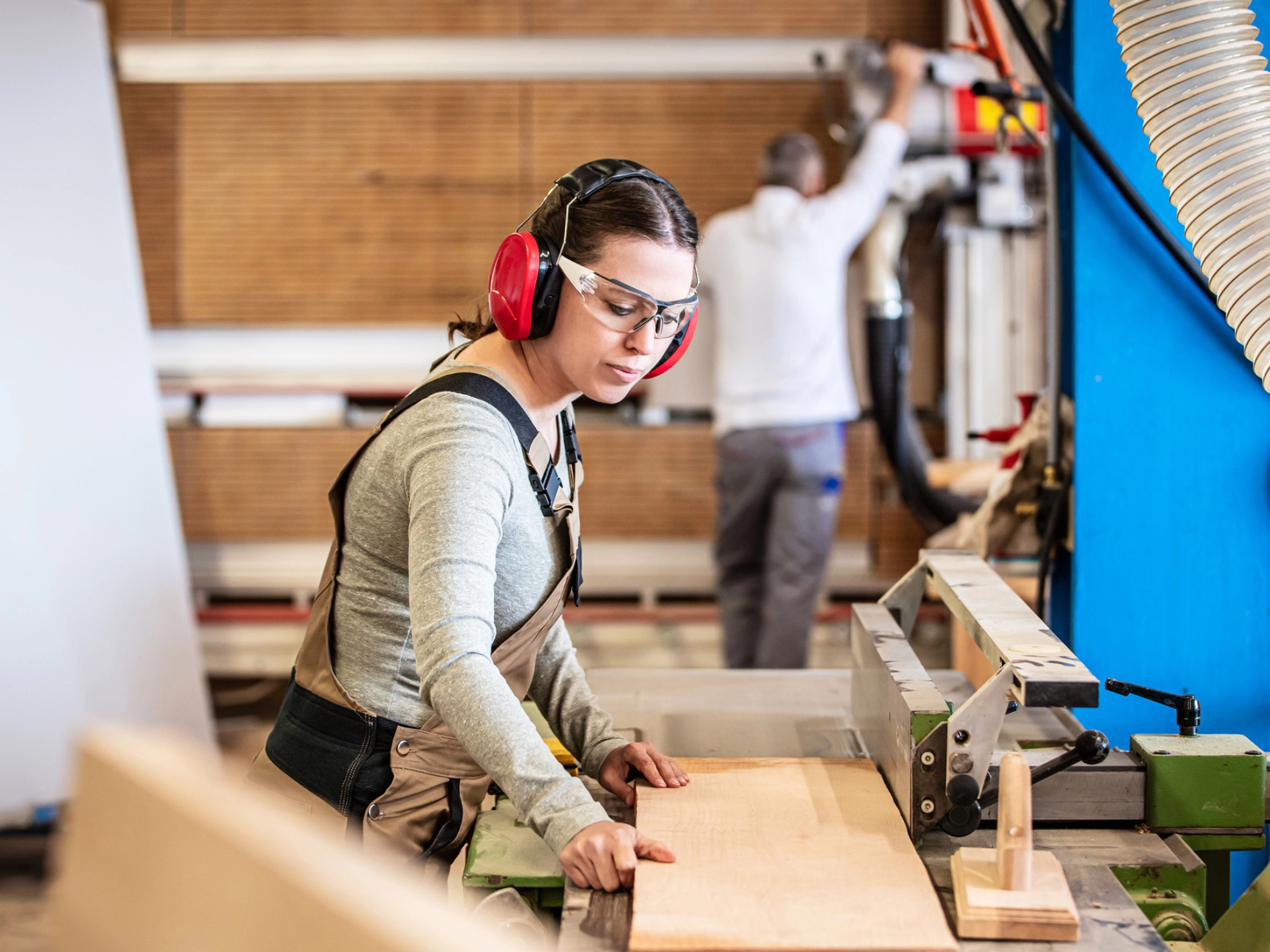Eine Schreinerin mit Gehörschutz und Schutzbrille arbeitet in einer Werkstatt mit Holz.