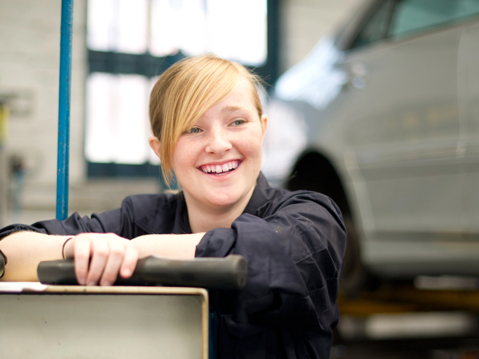 Eine junge blonde Frau mit einem Werkzeug in der Hand steht lächelnd in einer KFZ-Werkstatt.